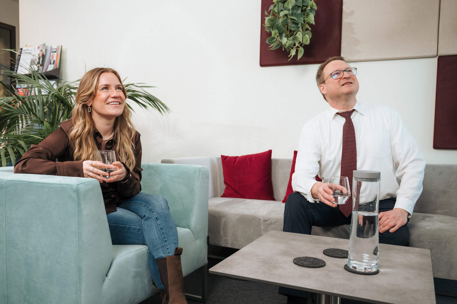 Zwei Personen sitzen in einem modernen Büro auf Sesseln und halten jeweils ein Glas Wasser